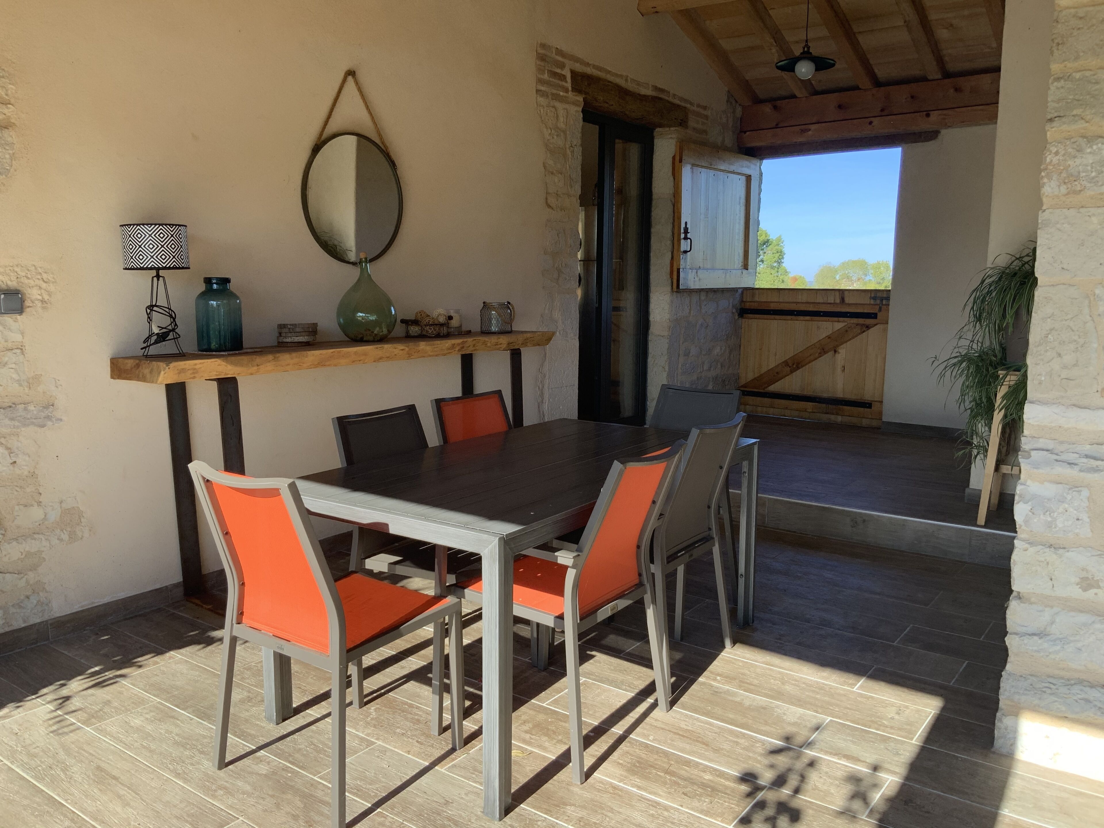 An outdoor dining area with a wooden table and chairs, a hanging mirror, and a shelf with decorative items.