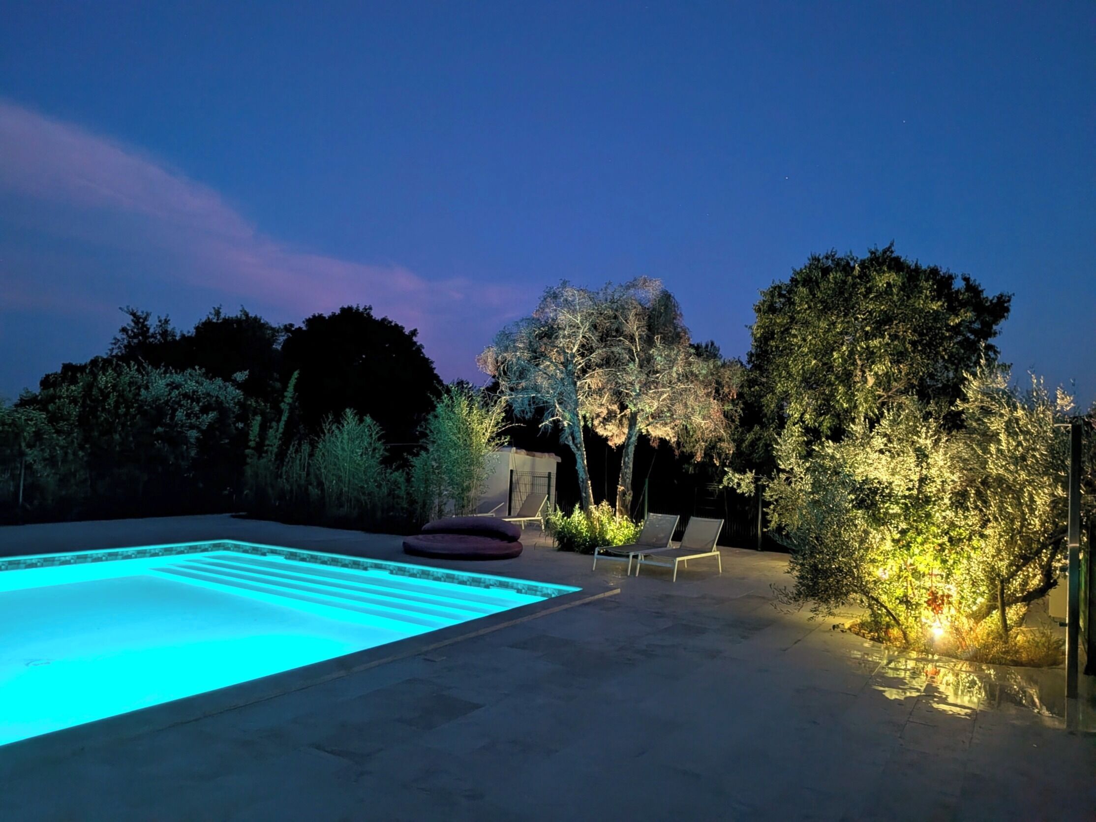 Ein Schwimmbecken mit Fliesenveranda, umgeben von Bäumen und einigen Liegestühlen.