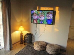 A living room with a television mounted on the wall, a lamp, a small table, and two ottomans. - In Uzès, 93m² airco+private terrace, sw.-pool 9X9, WIFI, huge garden, very quiet (Uzès)
