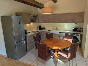 A modern kitchen with a stainless steel refrigerator, a built-in oven, and a coffee maker. There is a round wooden table with chairs and a hanging light fixture above. - In Uzès, 93m² airco+private terrace, sw.-pool 9X9, WIFI, huge garden, very quiet (Uzès)