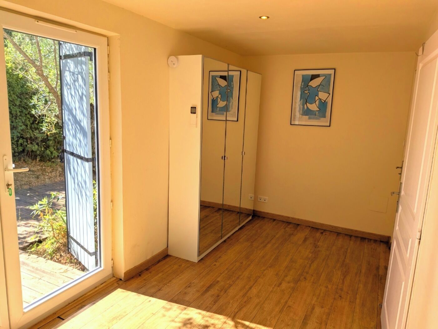 Une pièce dotée d’une porte-fenêtre coulissante donnant sur un jardin, d’une armoire blanche et de deux tableaux abstraits encadrés accrochés au mur.