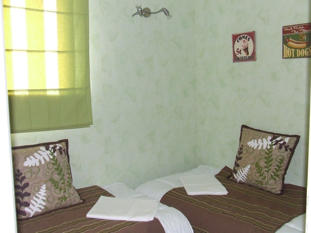 Ein Zimmer mit zwei Betten, jedes mit Kissen und Decke. An den Wänden befinden sich Verzierungen und ein Fenster mit Jalousien.