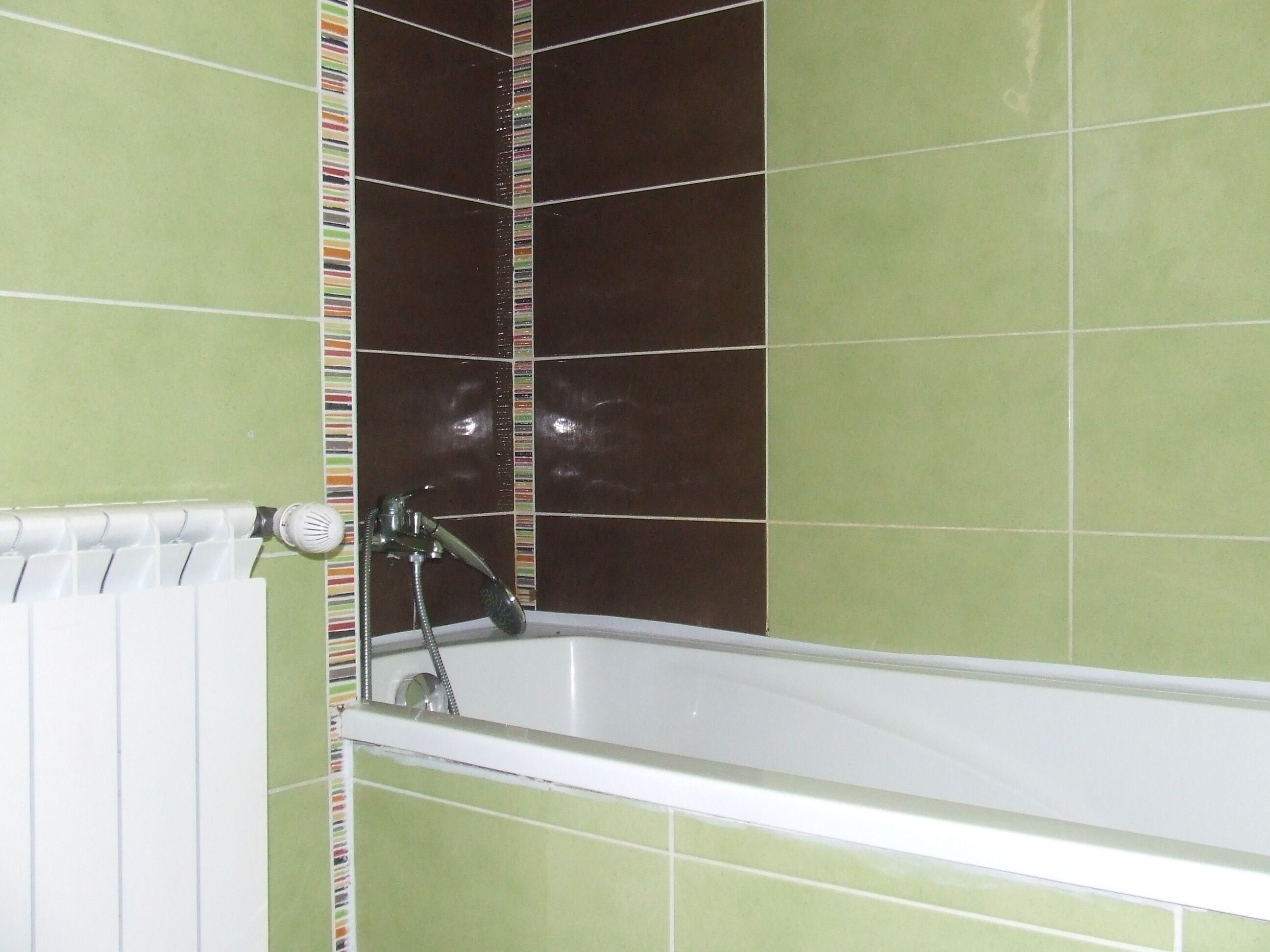 A bathroom with a bathtub, green and brown tiles, and a white radiator.