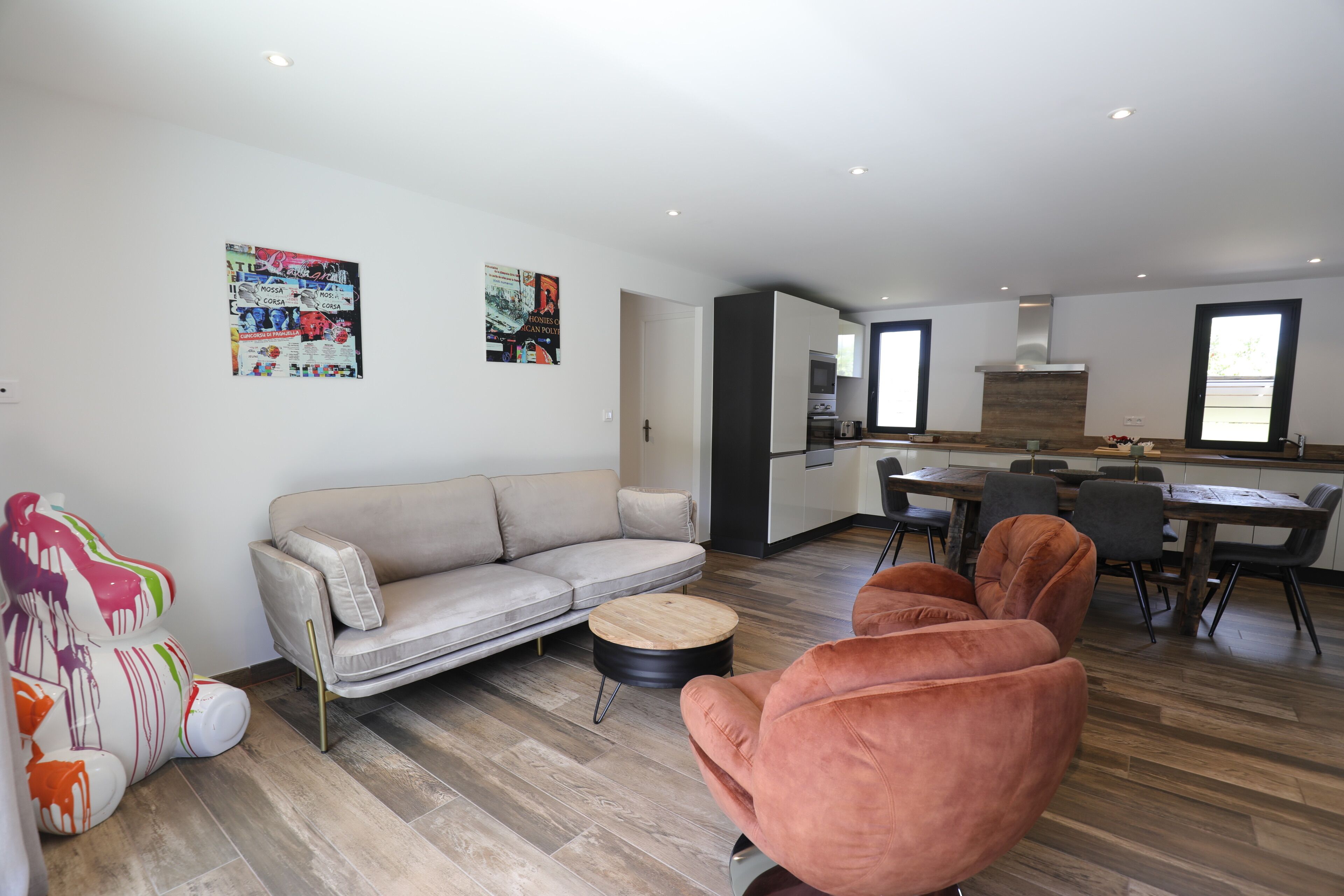 A modern living room with a sofa, a round coffee table, and a pink armchair. There is a kitchen area with a dining table and chairs visible in the background.
