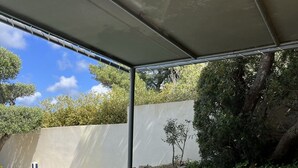 Un área de comedor al aire libre con una mesa de vidrio y sillas color naranja, rodeada por una piscina y follaje.