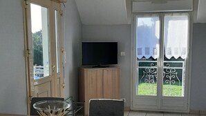 Ein Essbereich mit einem Holztisch, zwei Stühlen, einem Fernseher auf einer Holzkommode und einem Balkon mit Blick auf Grünflächen.