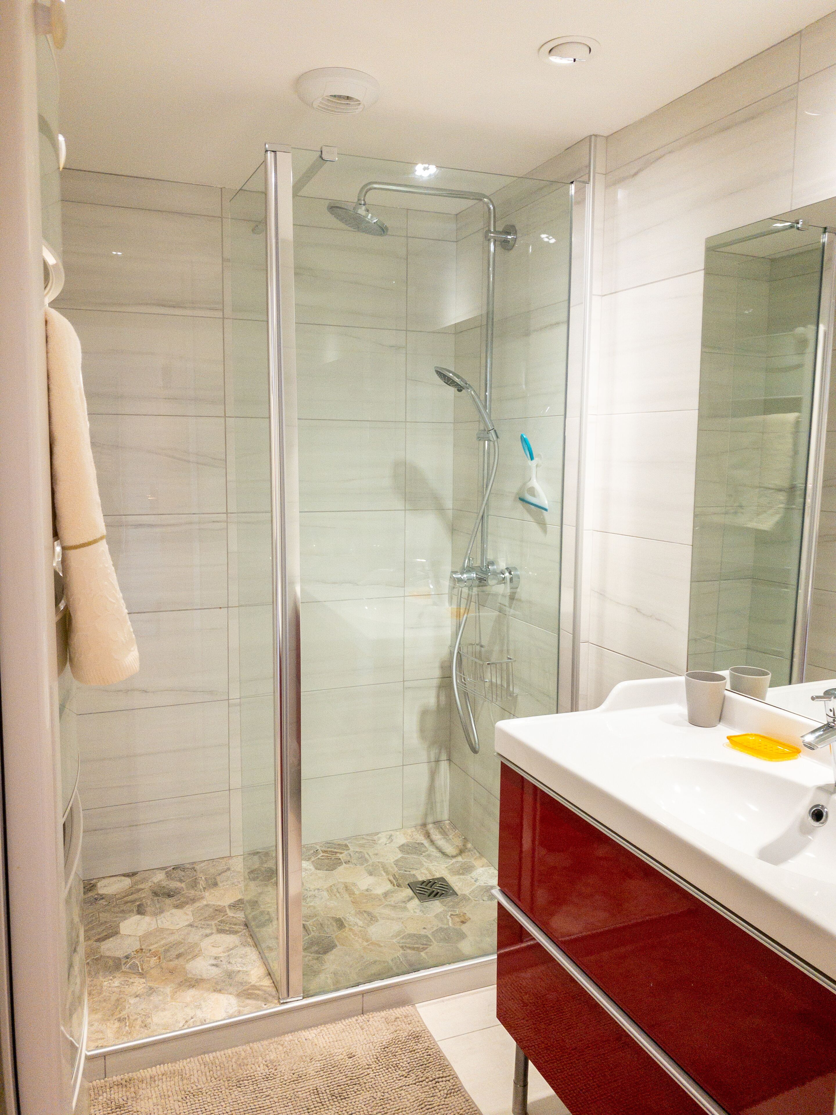 A modern bathroom with a glass-enclosed shower, a white sink with a red cabinet, and a mirror above the sink.