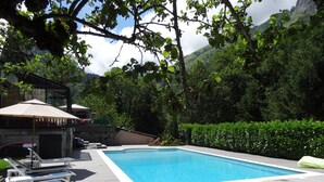 Una piscina con sillas de descanso y una zona cubierta, rodeada de vegetación y colinas.