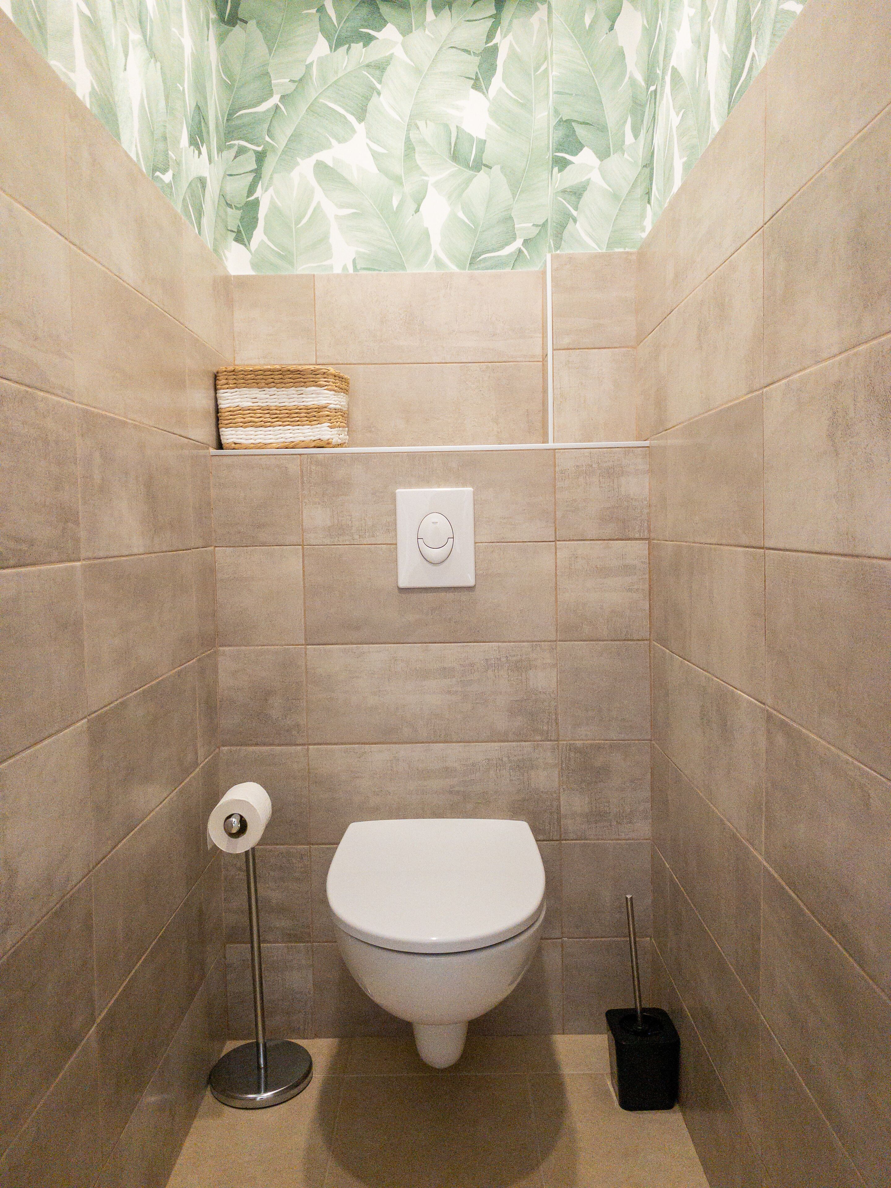 Une petite salle de bain carrelée, équipée de toilettes, d’un sèche-mains et d’une poubelle.