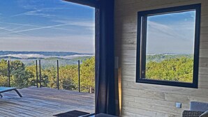 A room with a large window offering a view of a forested landscape. There is a wooden deck outside the window, and the room has wicker furniture and a table. - GRANGE AUX AMIS in SARLAT, an ecolodge with a nice view  of the DORDOGNE valley (SARLAT)