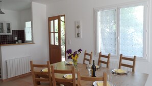 A dining area with a wooden table set for four, surrounded by chairs. A vase with flowers is on the table, and there is a bottle of wine. The room has white walls, a window with frosted glass, and a door leading outside. - Gîte La Salsepareille 6-person house near Pézenas with garden and air conditioning (Caux)