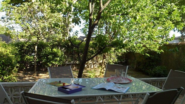 A glass table with chairs, a book, and a tray with glasses and a pitcher, set outdoors with a tree and garden in the background. - Gîte La Salsepareille 6-person house near Pézenas with garden and air conditioning (Caux)