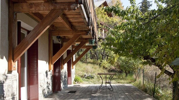 A wooden deck with a table, surrounded by stone walls and red shutters, leading to a covered porch area.
