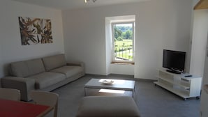 A living room with a sofa, a coffee table, a TV on a stand, and a window with a view of greenery. - HOUSE RENTAL IN CASTELJAU (Berrias-et-Casteljau)