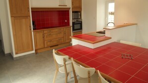 A kitchen with wooden cabinets, a red tiled backsplash, a built-in oven, and a dining area with a red table and white chairs. - HOUSE RENTAL IN CASTELJAU (Berrias-et-Casteljau)