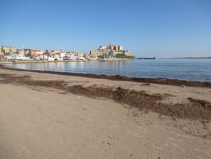 A coastal cityscape with a prominent castle-like structure on a hill, a sandy beach in the foreground, and a marina with boats.