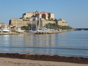 A coastal town with a prominent castle-like structure, a marina with several boats, and a sandy beach in the foreground. - Sea view, apartment with terrace in residence. Swimming pool, parking, Wifi, Air conditioning. (Lumio)