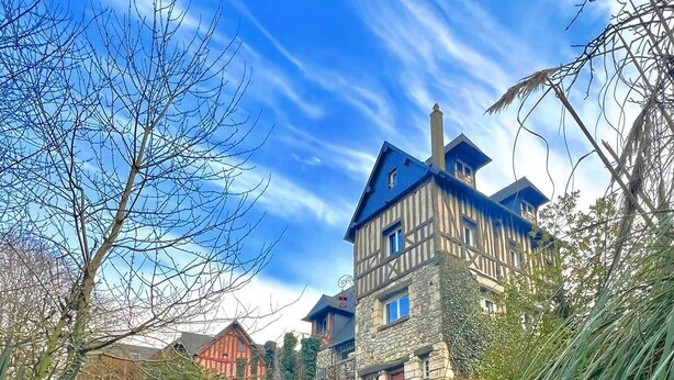 Un edificio grande de varios pisos con fachada de piedra y techo azul, rodeado por un jardín cuidado y un pequeño estanque.