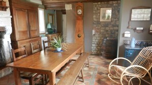 Un comedor rústico con una mesa de madera, bancos y sillas de mimbre. Se ve un reloj y una pared de piedra al fondo.