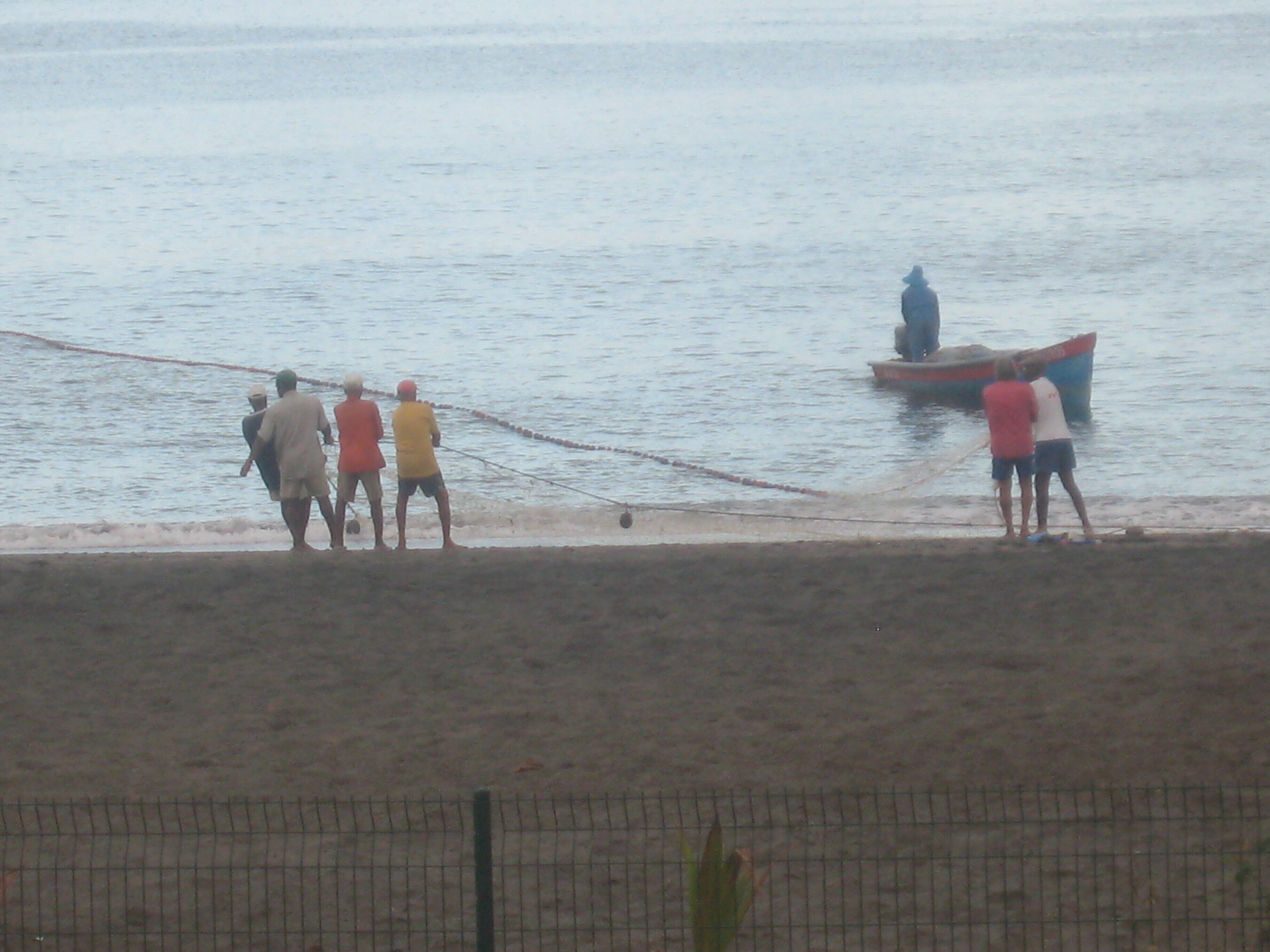 A group of people by the water, some standing and some sitting, with a boat in the background.