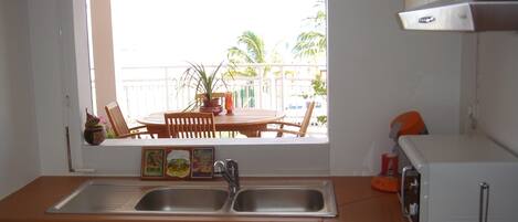 A kitchen with a double sink, wooden table, chairs, and a view of a balcony with plants and a railing.
