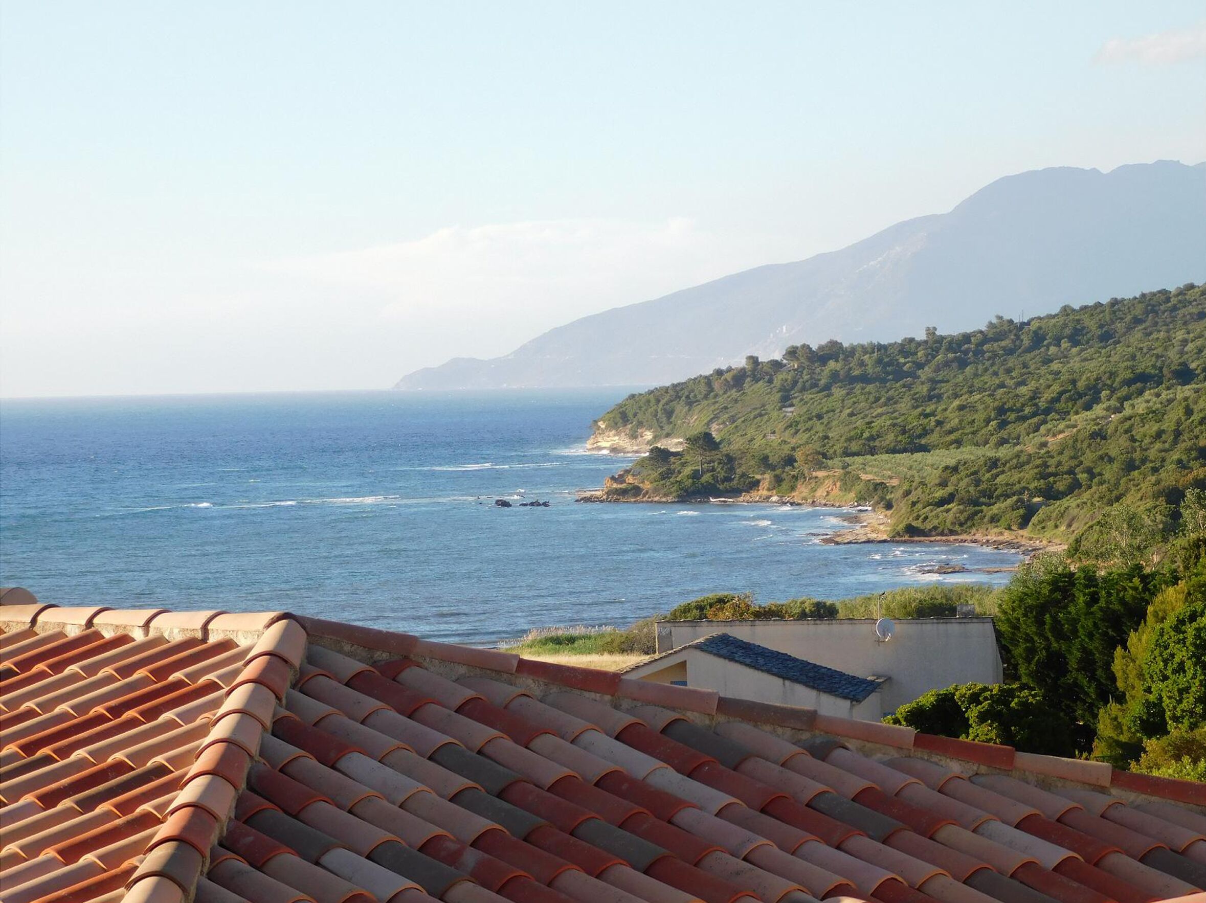 A coastal view with a tiled roof, a clear sky, and a mountainous backdrop.