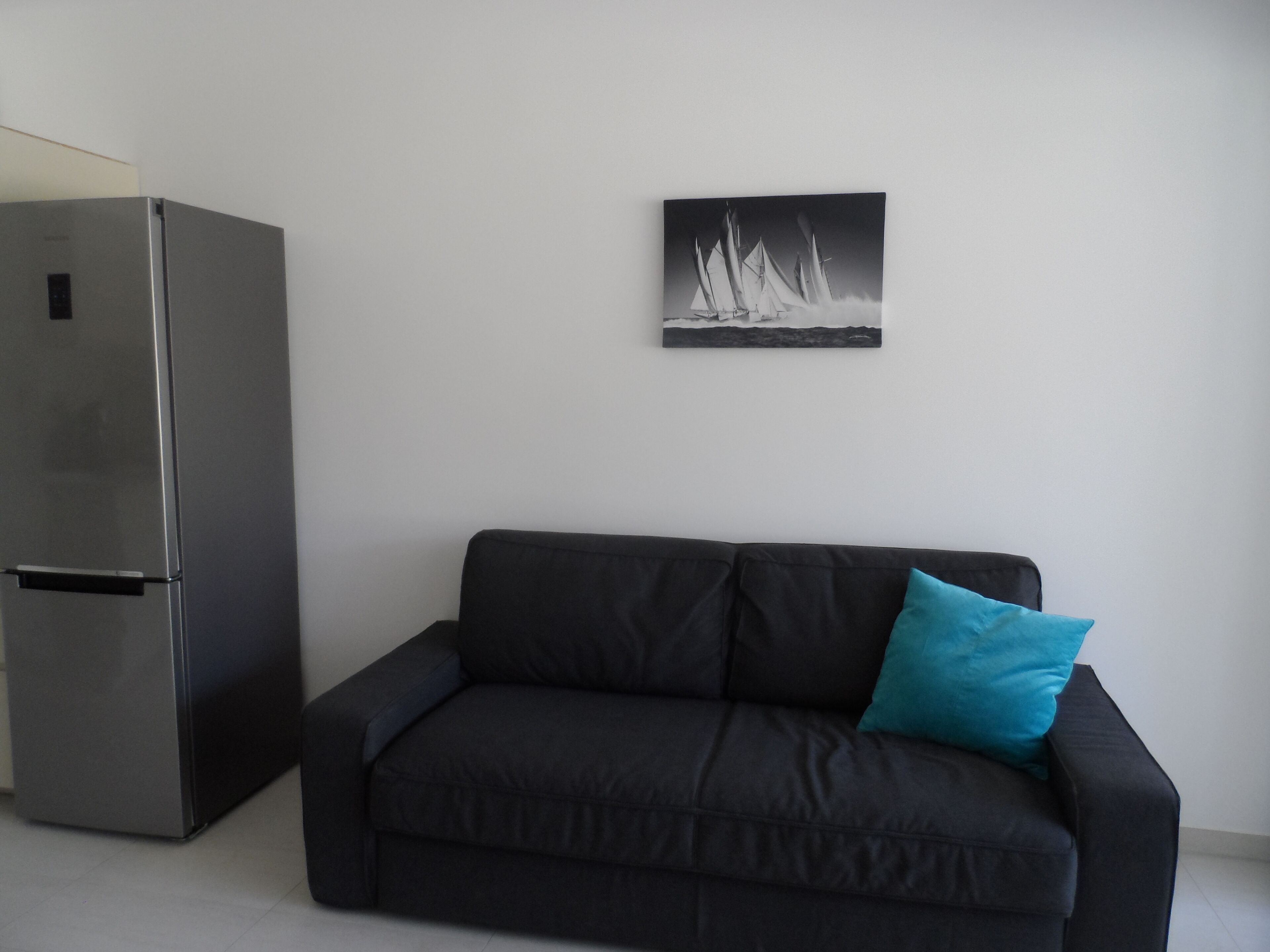 A modern living room with a black sofa and a teal pillow, a stainless steel refrigerator, and a black and white sailboat painting on the wall.