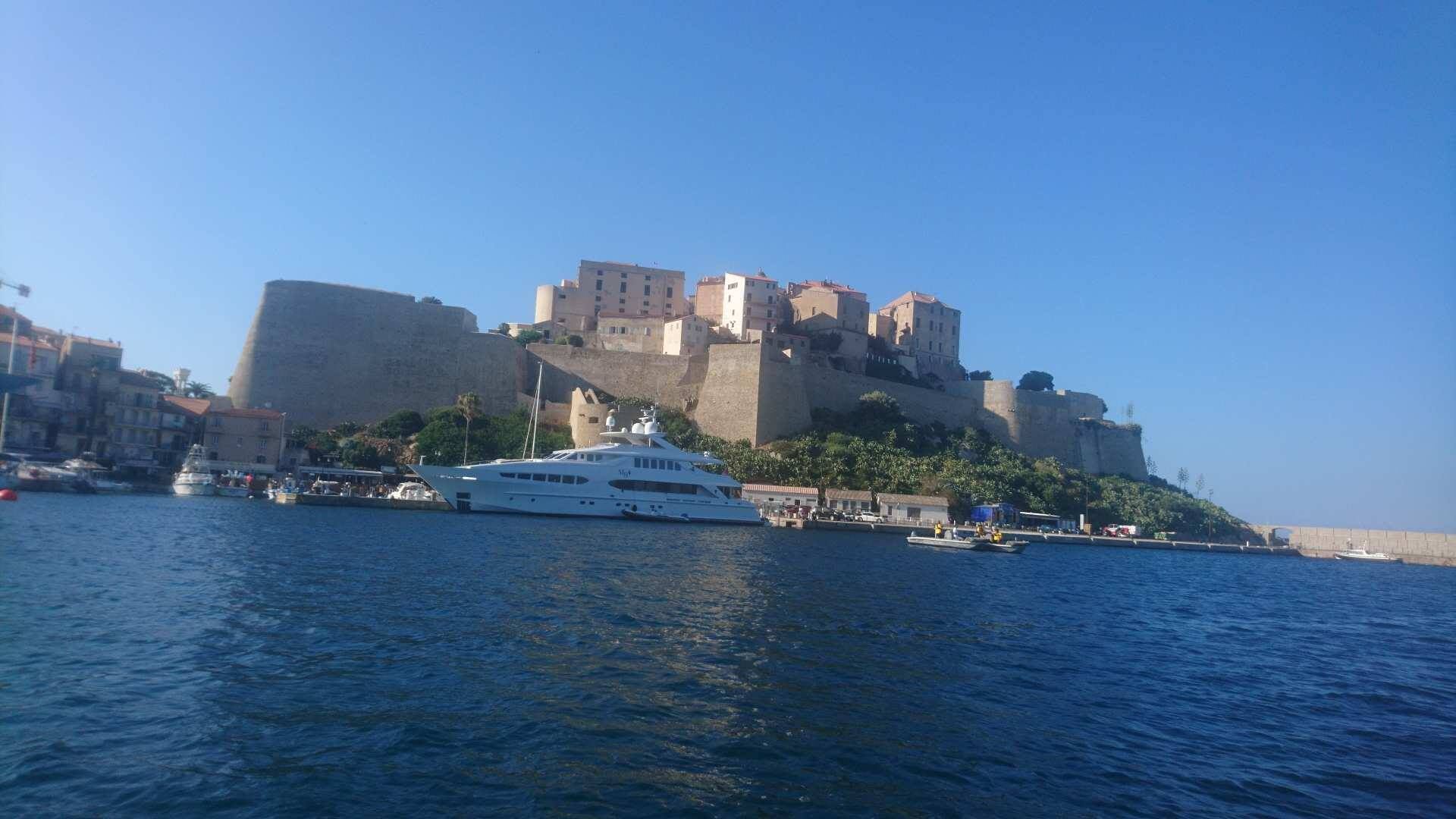 Un grand château perché sur une colline, dominant un plan d’eau, avec un yacht amarré à proximité.
