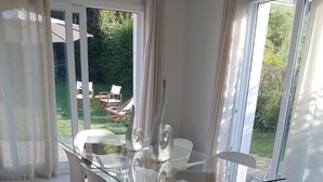 A modern dining area with a glass table and white chairs, large windows with curtains, and a view of a garden.