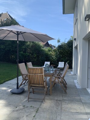 Une terrasse extérieure avec une table et des chaises, un grand parasol et une vue sur une maison en arrière-plan.