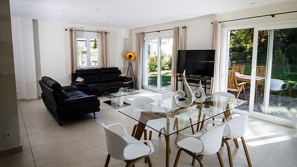 A modern living room with a glass dining table, white chairs, a black sofa, and a TV.
