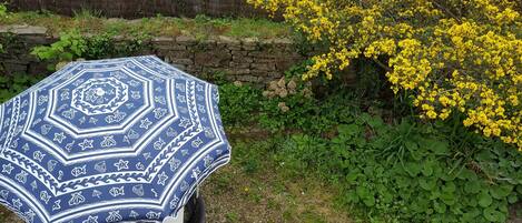 A blue patterned umbrella over a chair, a stone wall, and yellow flowering plants.