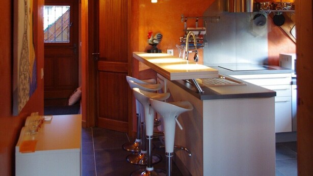 A rustic kitchen with wooden beams, a central island with bar stools, a hanging clock, and a window with a view.