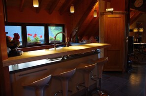 A kitchen with a bar area, wooden ceiling beams, and a window with flowers. - Charming gite with large garden 15 minutes from Kaysersberg (ALSACE)