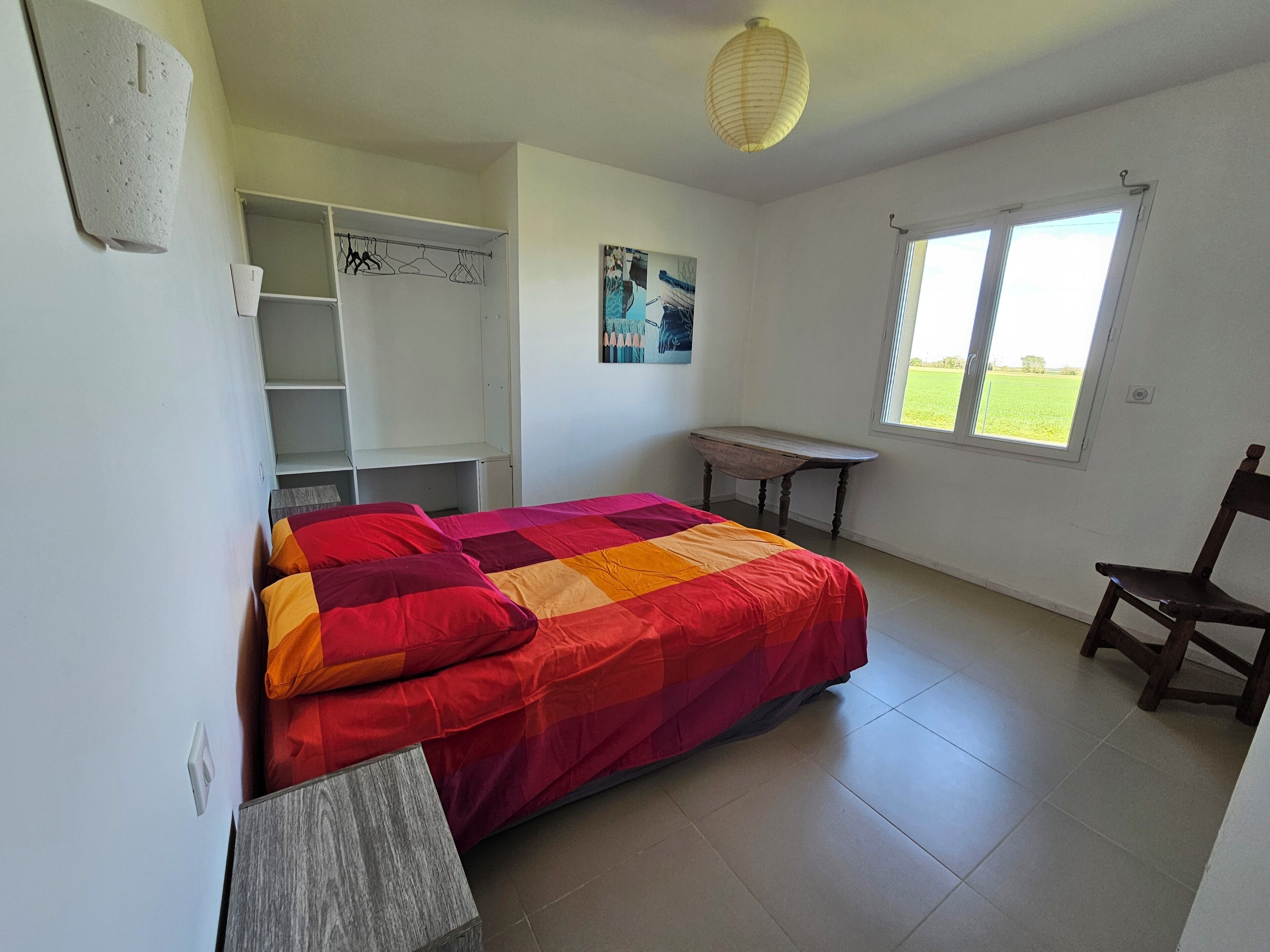 A bedroom with a bed, a desk, a chair, a wardrobe, and a window with a view of a green field.