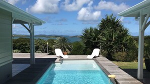 Una piscina rectangular con dos sillas de descanso blancas, rodeada por una terraza de madera y pérgolas blancas, con vistas a un paisaje pintoresco con árboles y una masa de agua.