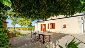 A rustic outdoor dining area with a round table and chairs, surrounded by greenery and a building with white walls and wooden shutters. - Charming cottage and nature, 18 ha, Périgord Noir-Dordogne -Lot -PISCINE and private SPA (Orliaguet)