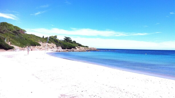 Ein Strand mit weißem Sand, klarem, blauem Wasser und einer hügeligen Landschaft im Hintergrund.