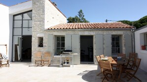 Een gebouw van steen met een pannendak, een patio met houten stoelen en een tafel, en een groot raam.