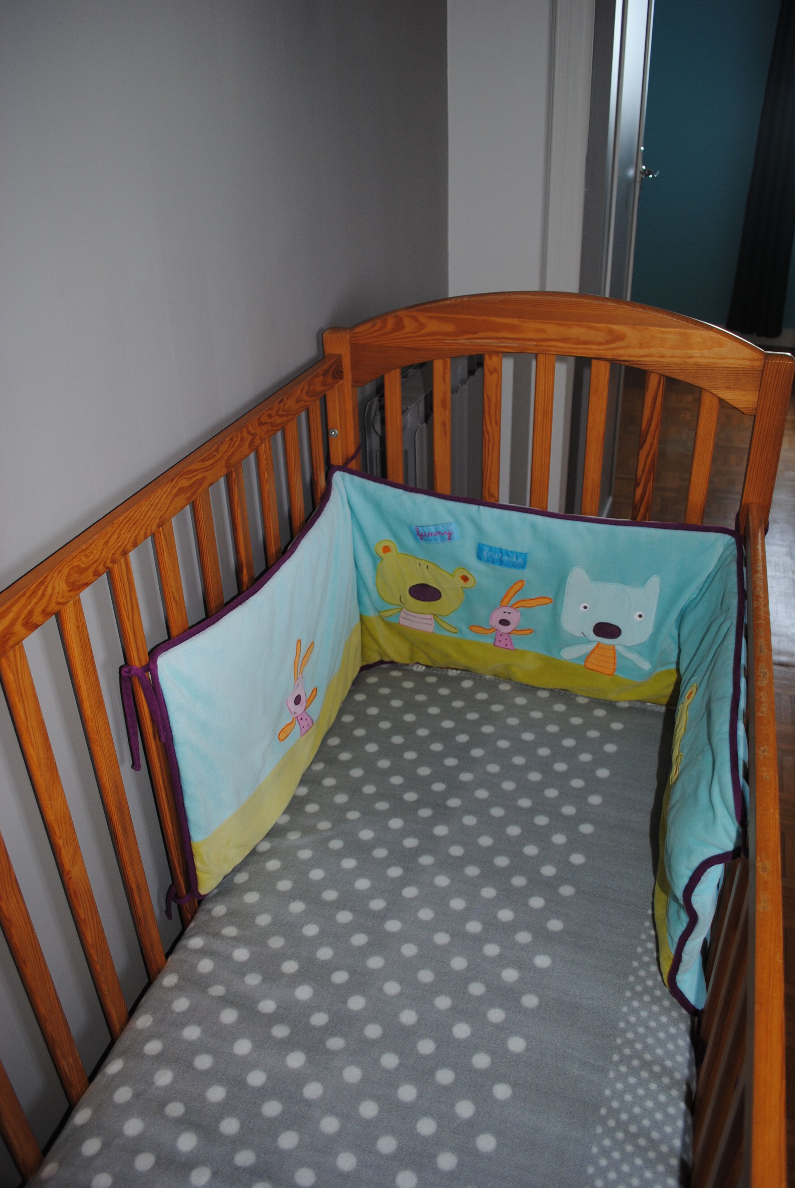 A wooden crib with a polka dot mattress and a colorful safety cover featuring cartoon characters.