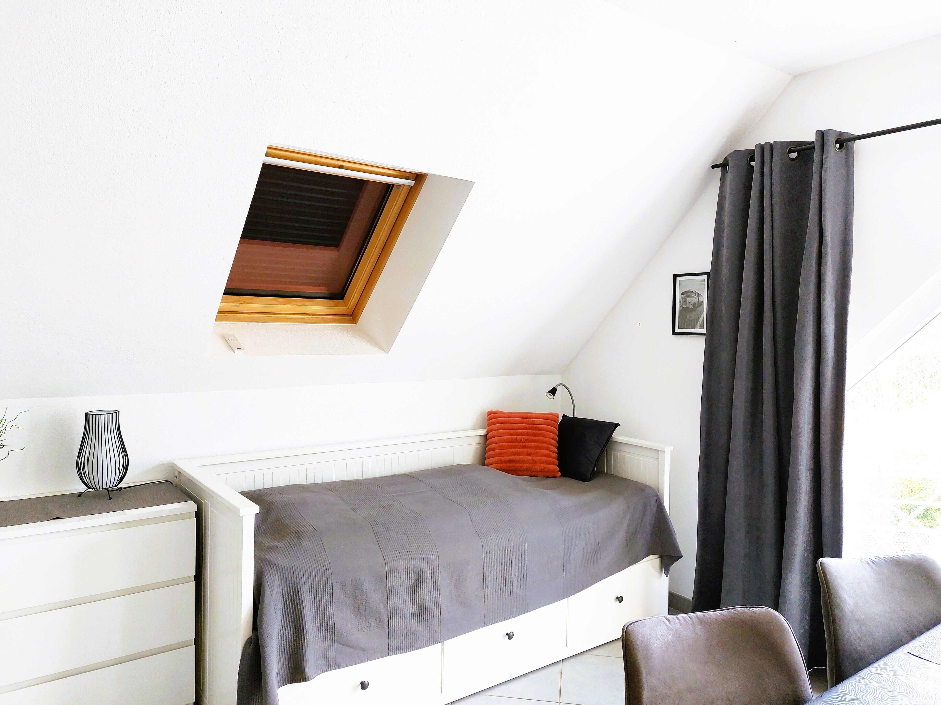 Ein Schlafzimmer mit schrägverdeckter Decke, einem Oberlicht, einem Bett mit grauer Bettdecke, zwei Kissen, einer weißen Kommode und einem Stuhl.