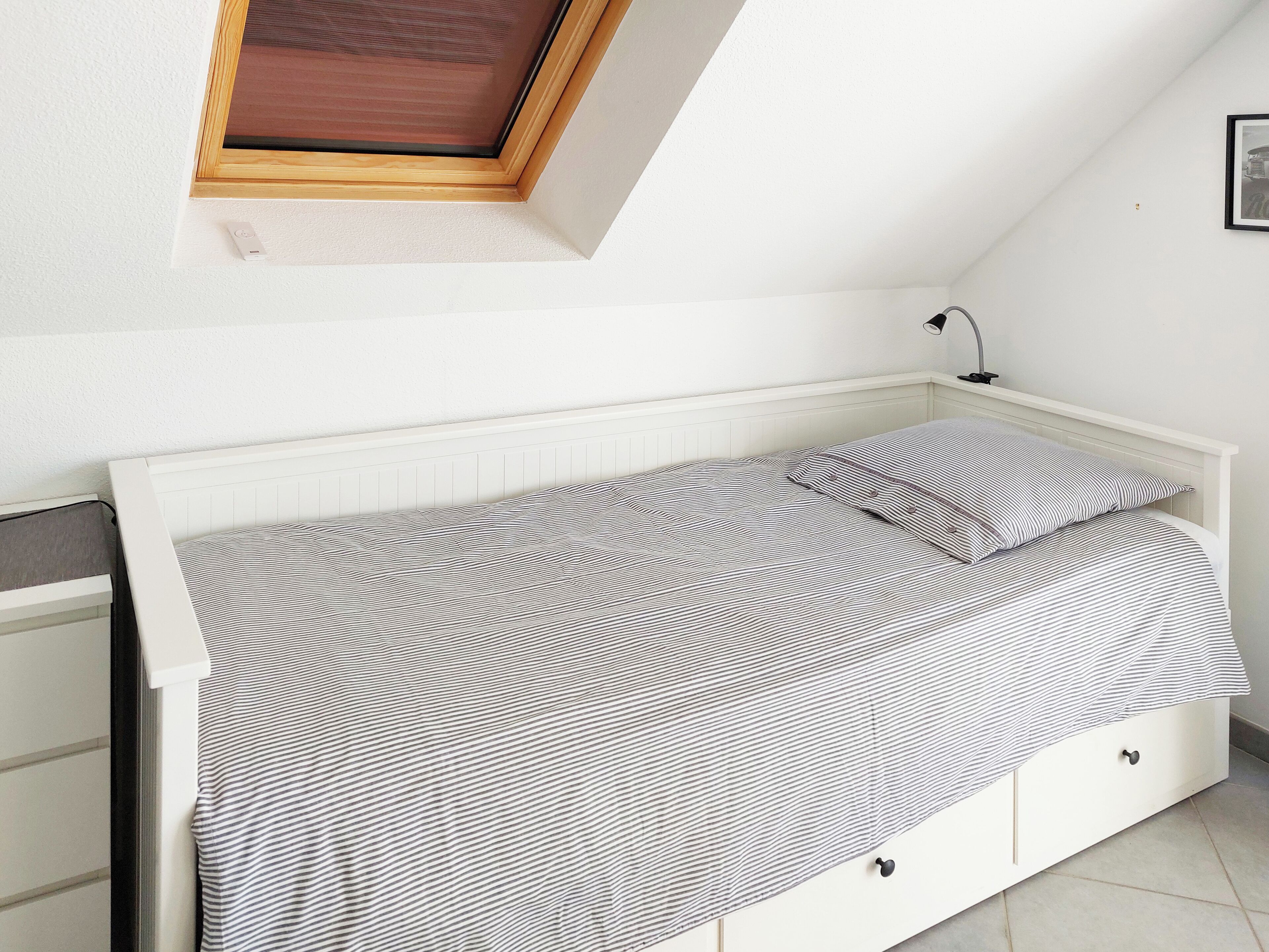 Ein kleines, ordentlich bezogenes Bett mit einer gestreiften Tagesdecke in einem Raum mit weißen Wänden und einer schrägen Decke.