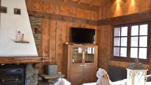 A cozy cabin interior with a stone fireplace, wooden walls, a television mounted above a wooden cabinet, and a large window letting in natural light. - Chalet with 4 bedrooms - 8 people - (Ideal Family, Group of friends) (Les Contamines-Montjoie)