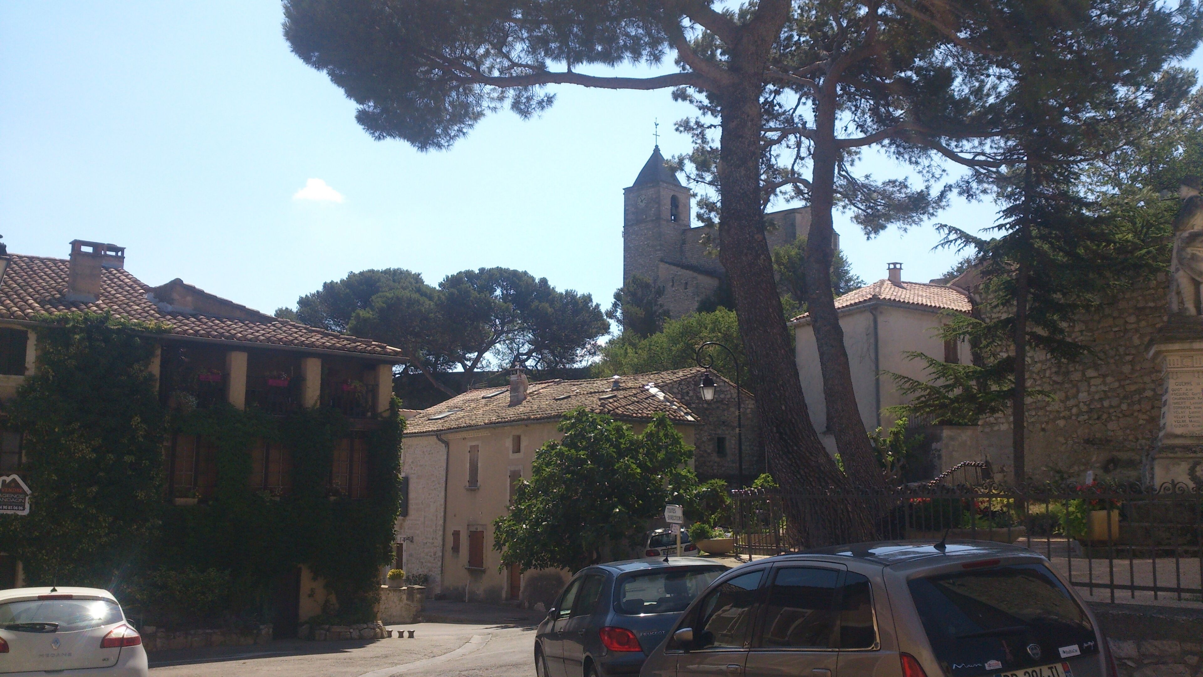 Une rue bordée d  ’immeubles, avec un clocher d’église et des voitures garées.