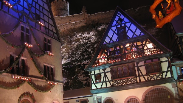 A traditional half-timbered building with a balcony, decorated with lights and garlands, and a clock tower in the background.