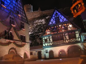 A traditional half-timbered building with a balcony, decorated with lights and garlands, and a clock tower in the background.