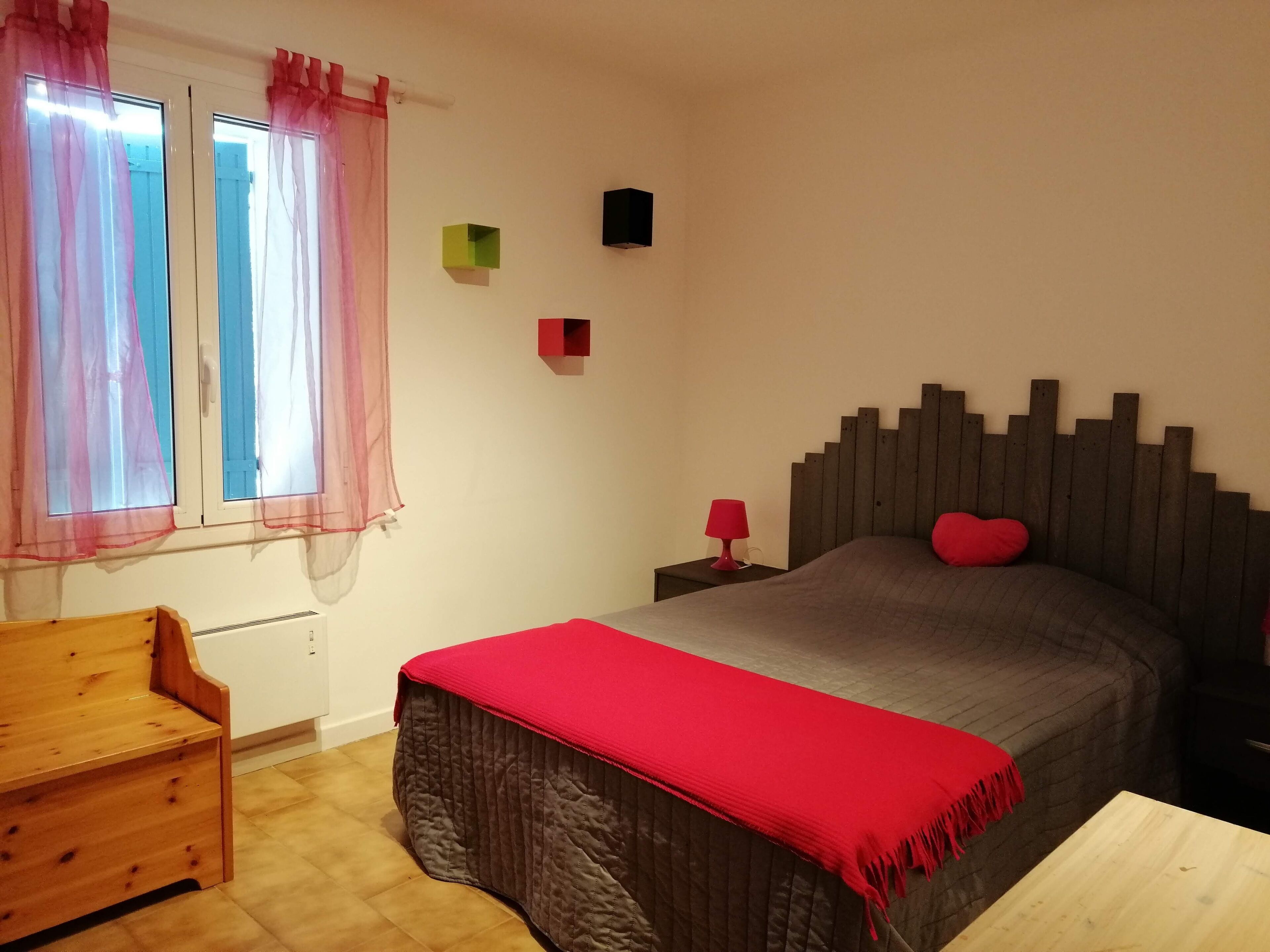 Un dormitorio con cama, un banco de madera, una mesita de noche y una ventana con cortinas traslúcidas.