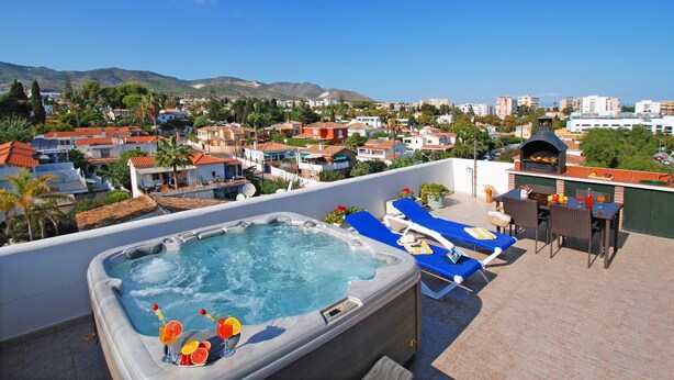 Terraza en la azotea con jacuzzi, sillas de descanso y zona de barbacoa, con vistas a una zona residencial con casas y montañas.