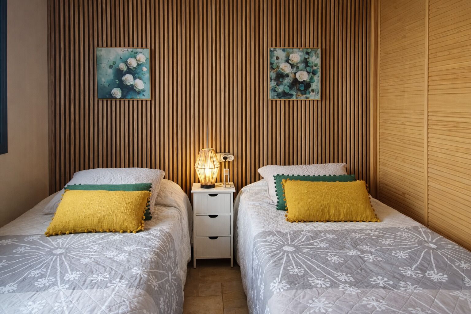 A room with two beds, each with a white floral bedspread and a yellow pillow. There are two wall-mounted paintings of flowers, a bedside lamp, and a white nightstand with drawers.