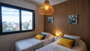 Un dormitorio con dos camas, una ventana con vista a la ciudad, una lámpara colgante y una lámpara de pared.